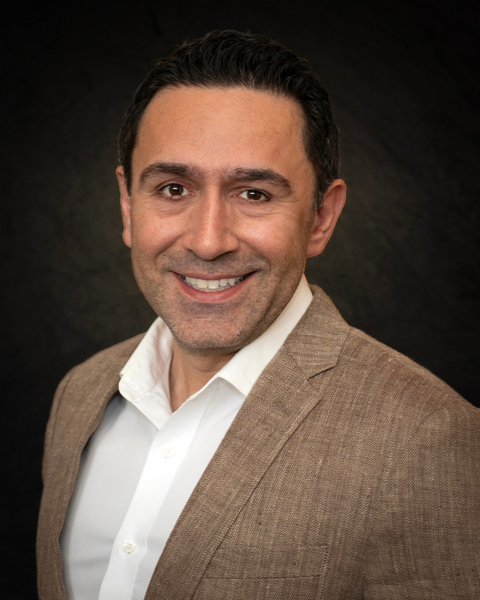 Portrait of a man with short dark hair wearing a white shirt and tan blazer, smiling against a dark background.
