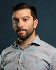 Portrait of a man with short dark hair and a beard wearing a gray button-up shirt, posed against a blue background.